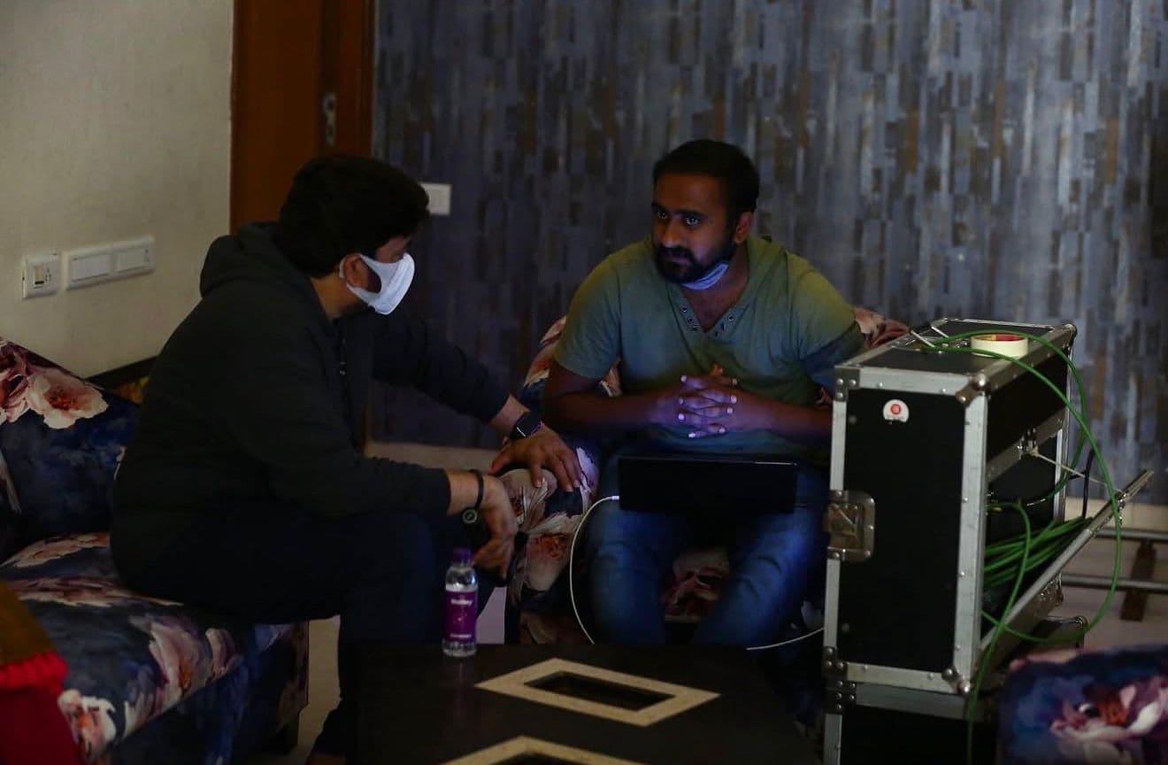 Two crew members on a floral sofa, one offering a water bottle to the other who is working on a laptop, camera case beside them
