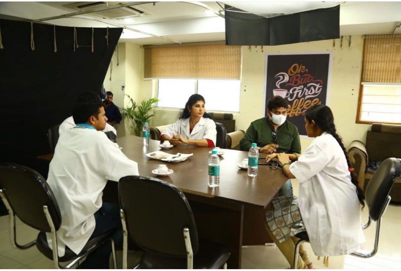 Five cast members in white coats seated around a table with tea, hospital-set interior