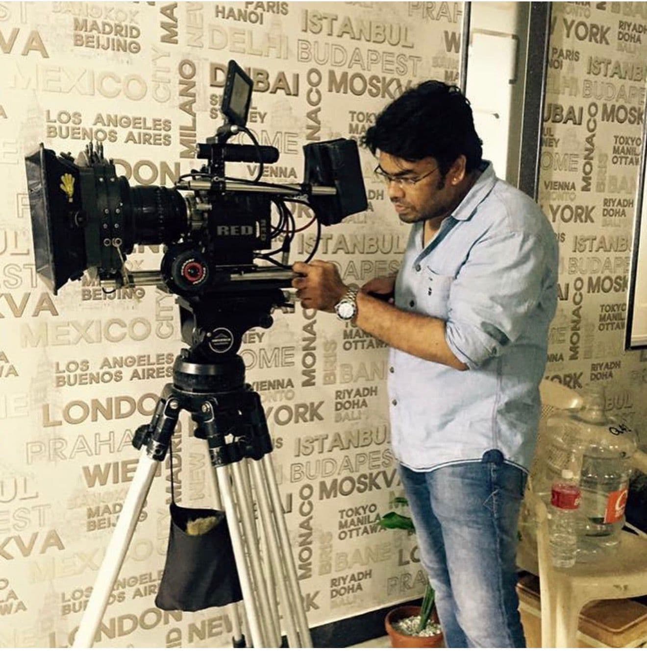 A crew member operating a RED cinema camera on a tripod against a wall lettered with world city names