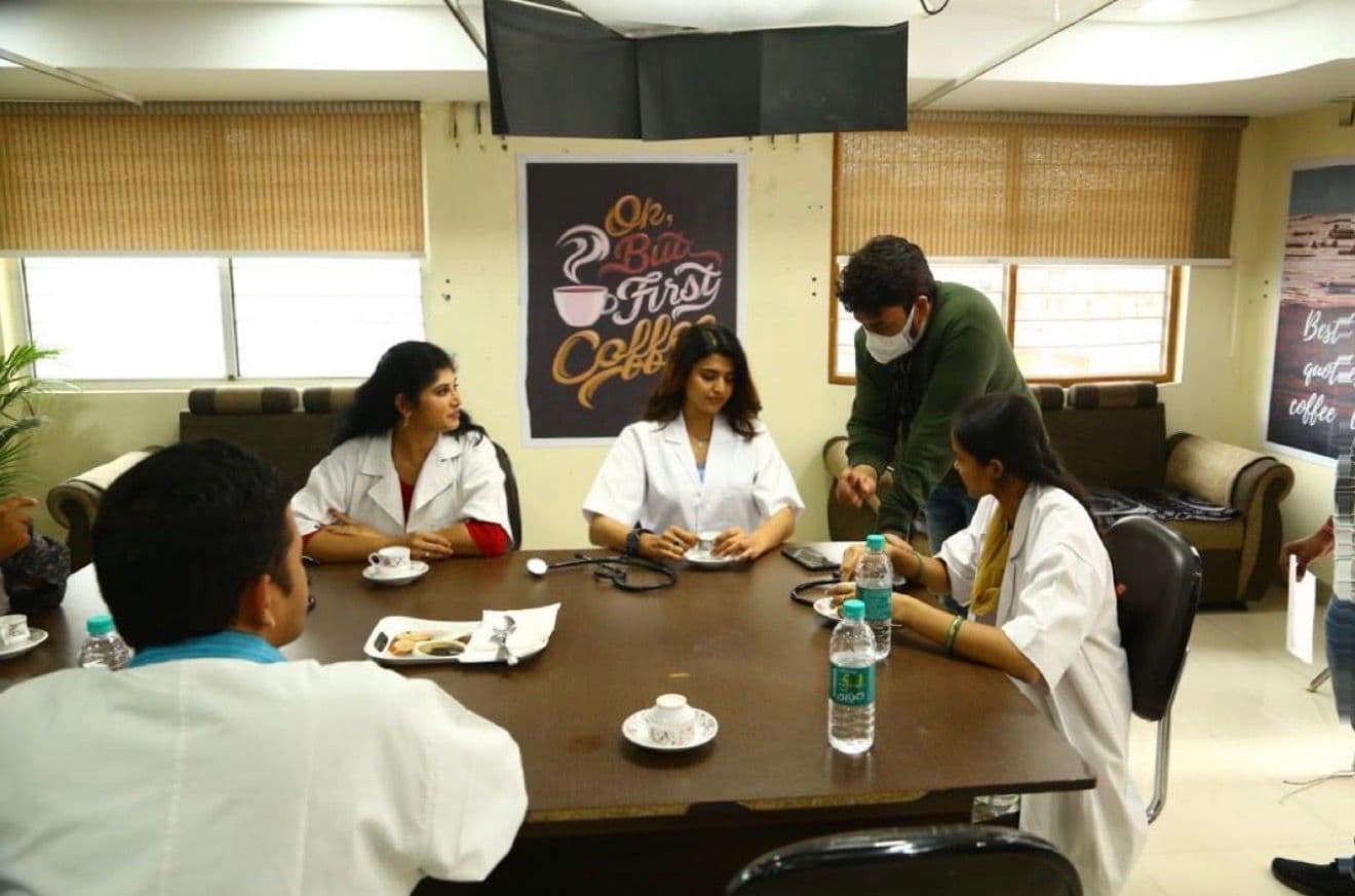 Cast in white coats around the hospital-set table, one standing and gesturing toward the group
