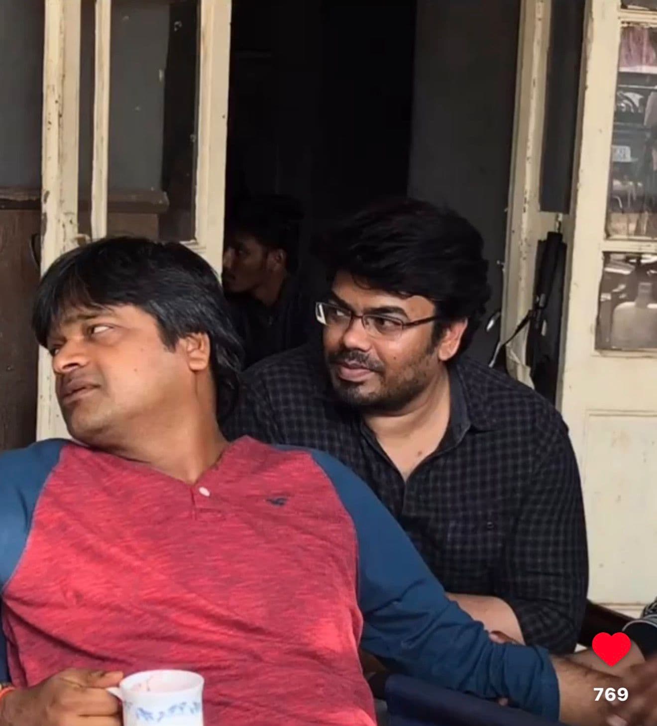 Two men seated together at an outdoor tea stall, one drinking from a mug