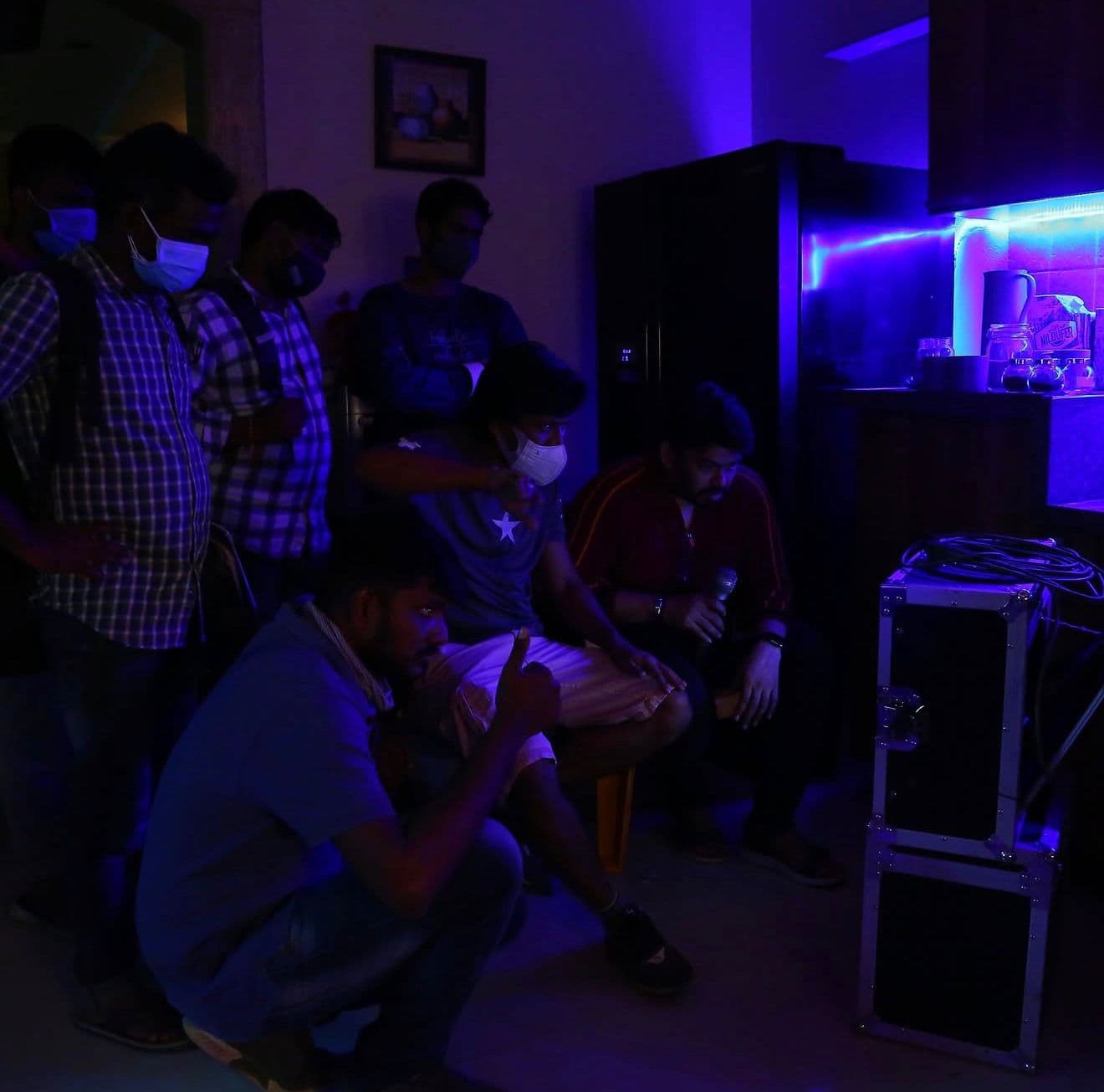 Cast and crew in face masks gathered around film equipment cases in a room lit with blue light