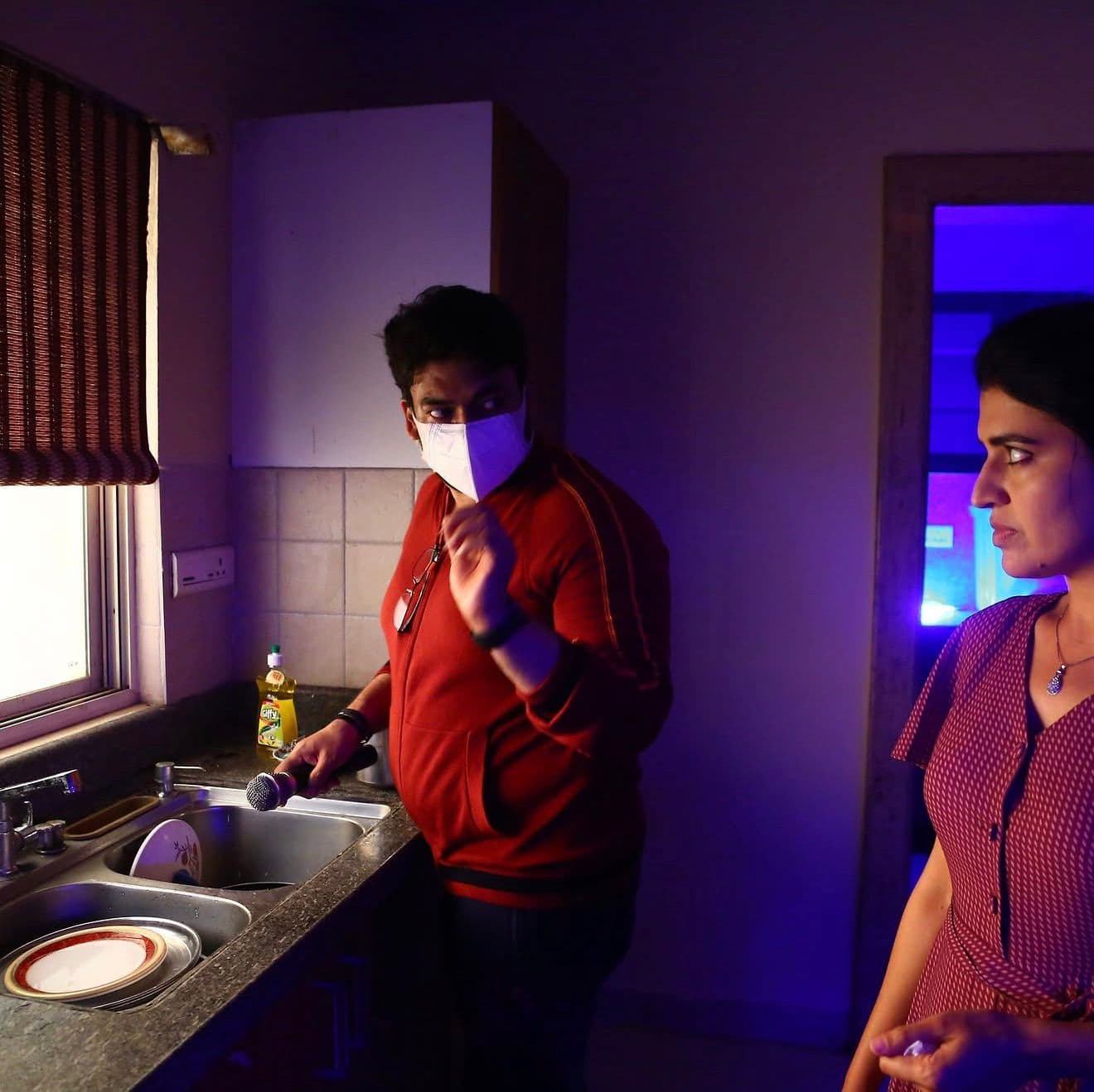 An actor in a red jacket and face mask gesturing in a kitchen, purple-lit interior