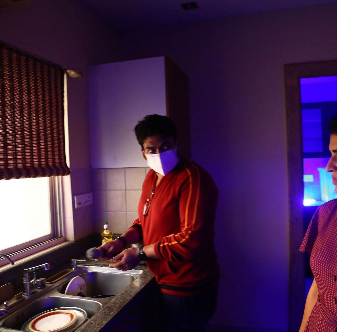 The same kitchen scene, the actor adjusting his mask beside the sink