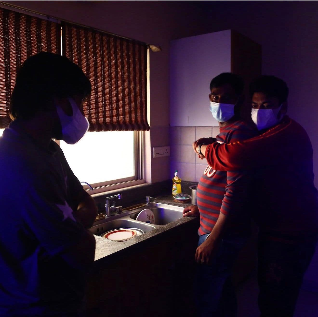 Two masked cast members embracing near the kitchen sink, a third figure watching in silhouette
