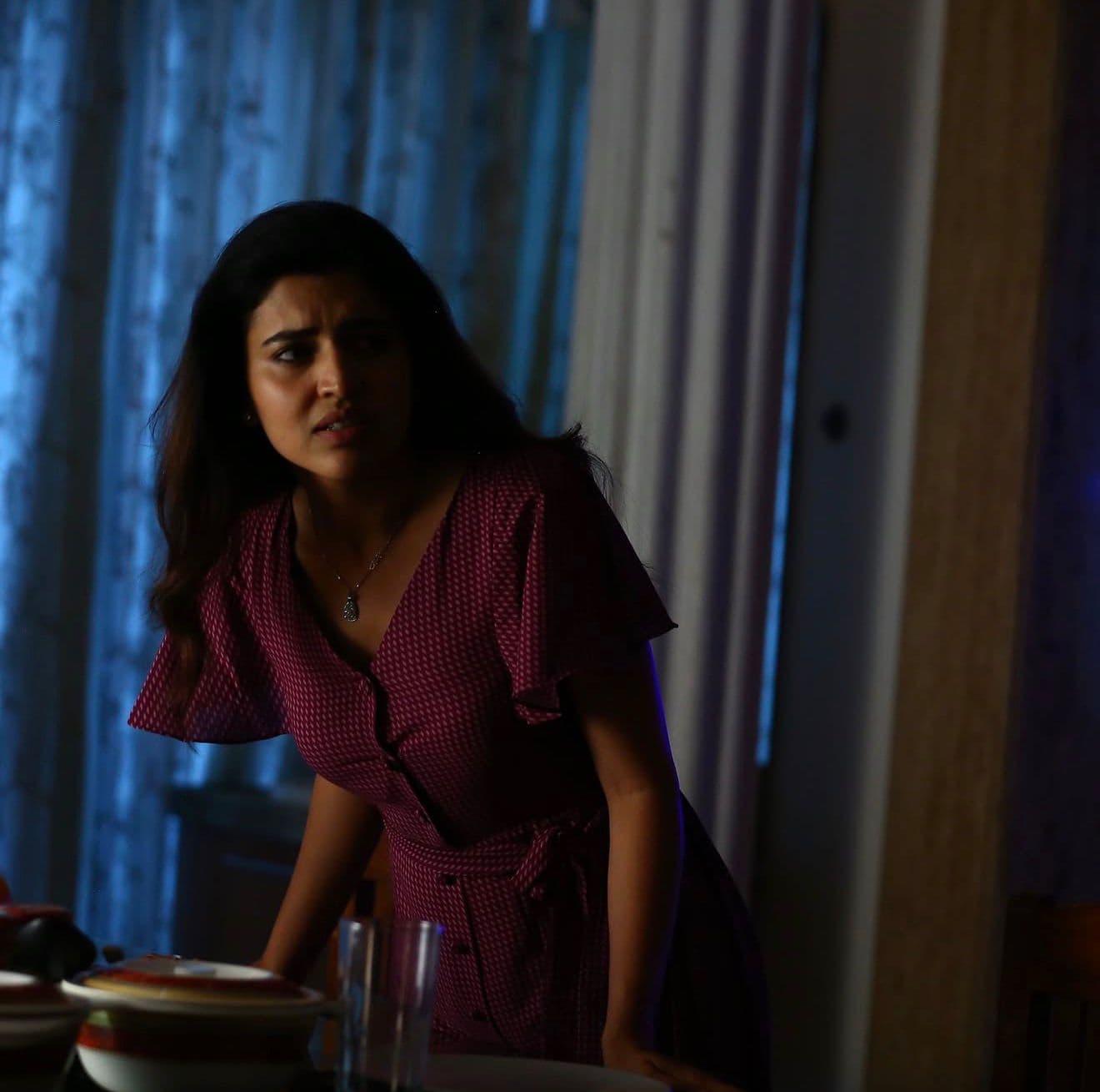 An actress standing at a dining table, backlit by curtains, dinnerware in the foreground