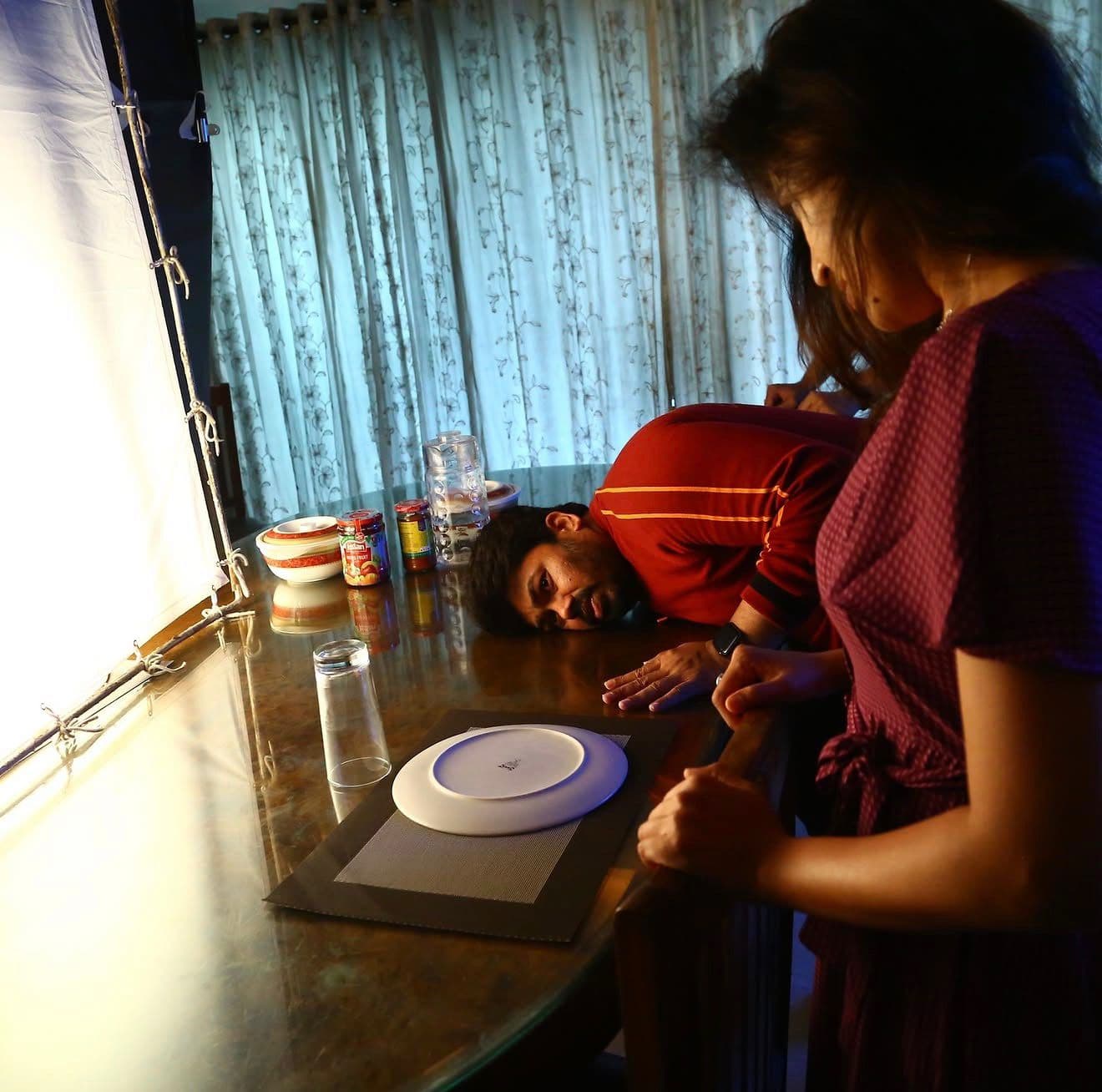 An actor lying face-down on a dining table beside an actress, curtained window behind them