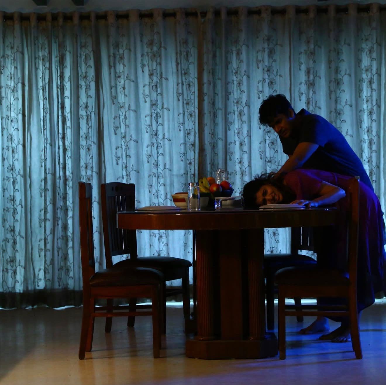 Two actors at a dining table in moody blue lighting, one leaning over the other who is lying on the table
