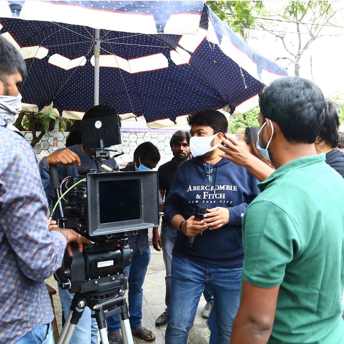 A crew member speaking into a microphone beside a camera rig under a large umbrella, outdoors
