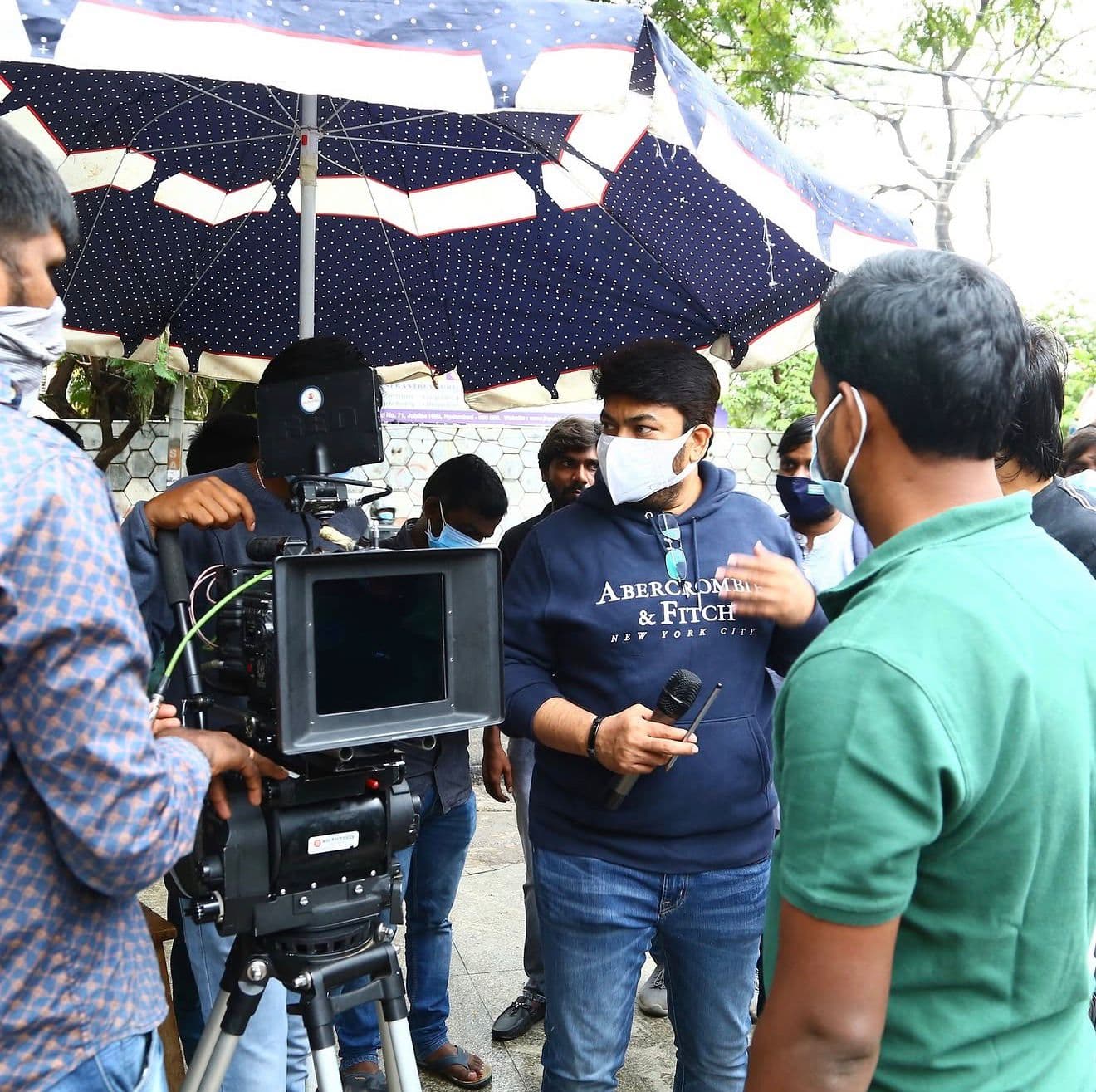 A crew member speaking with a microphone under an umbrella rig, cast and crew gathered around outdoors