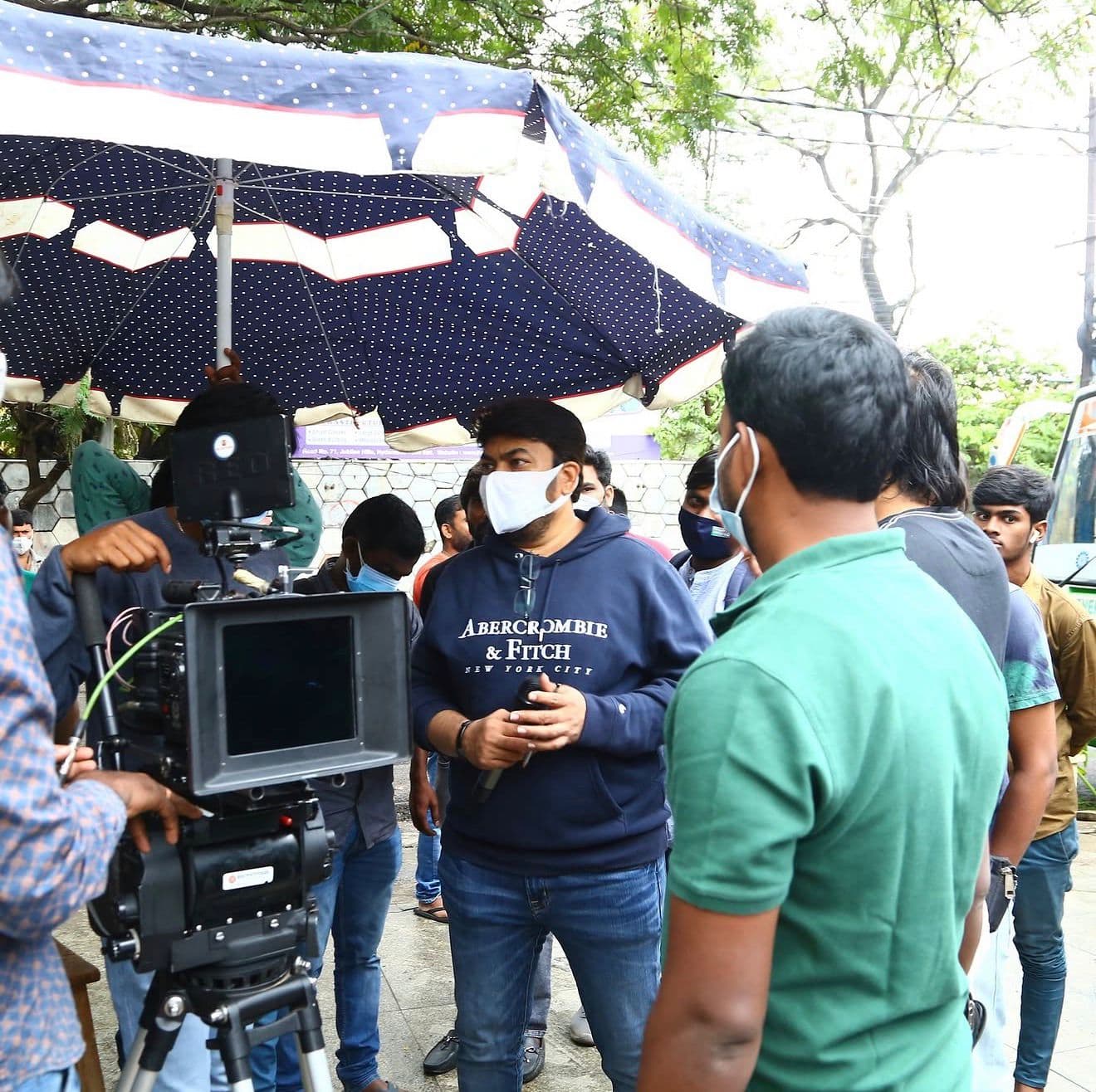 A crew member mid-conversation under an outdoor umbrella rig with cast and crew nearby
