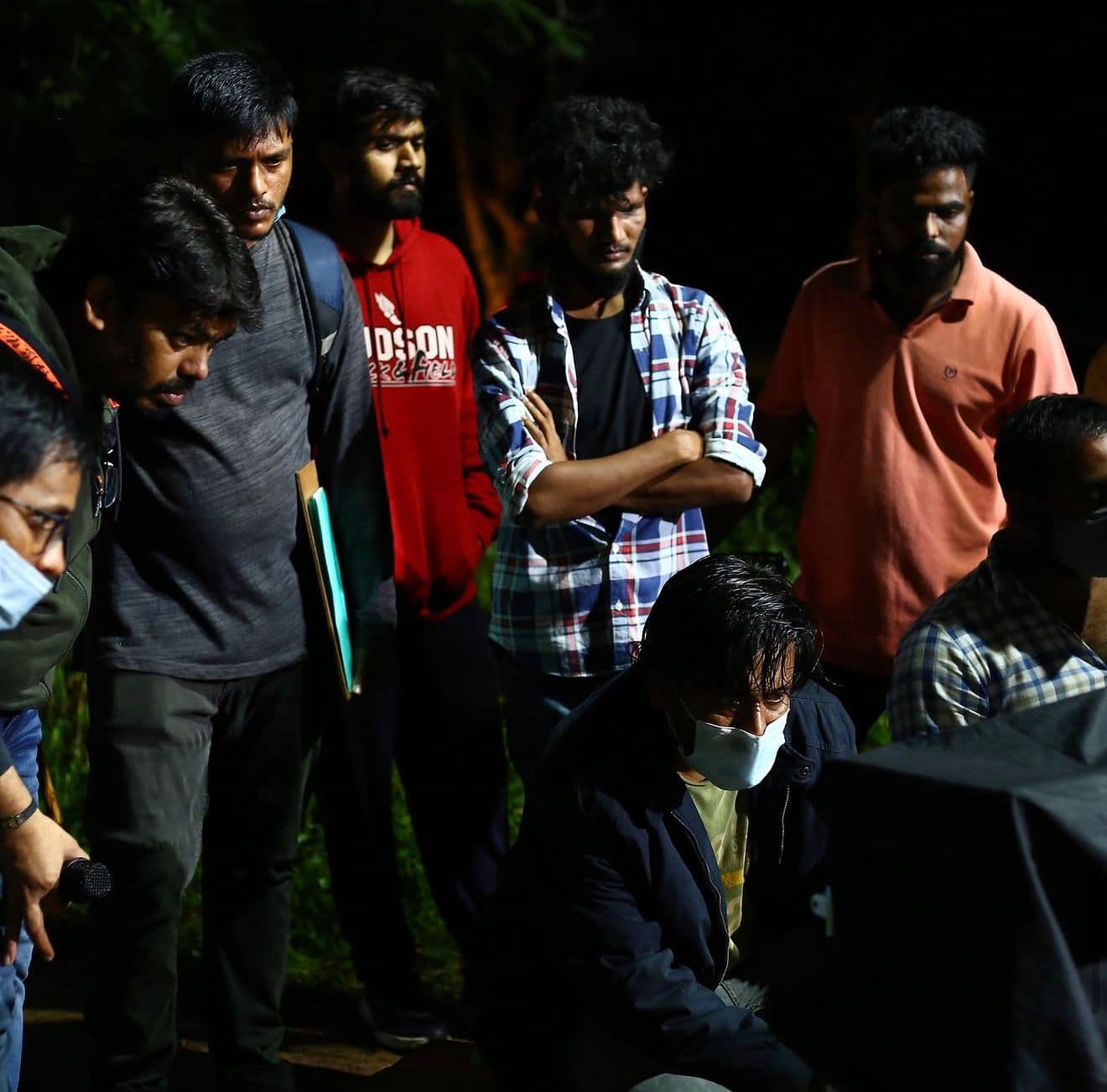 A crew group standing together outdoors at night, one crouched in a face mask in the foreground