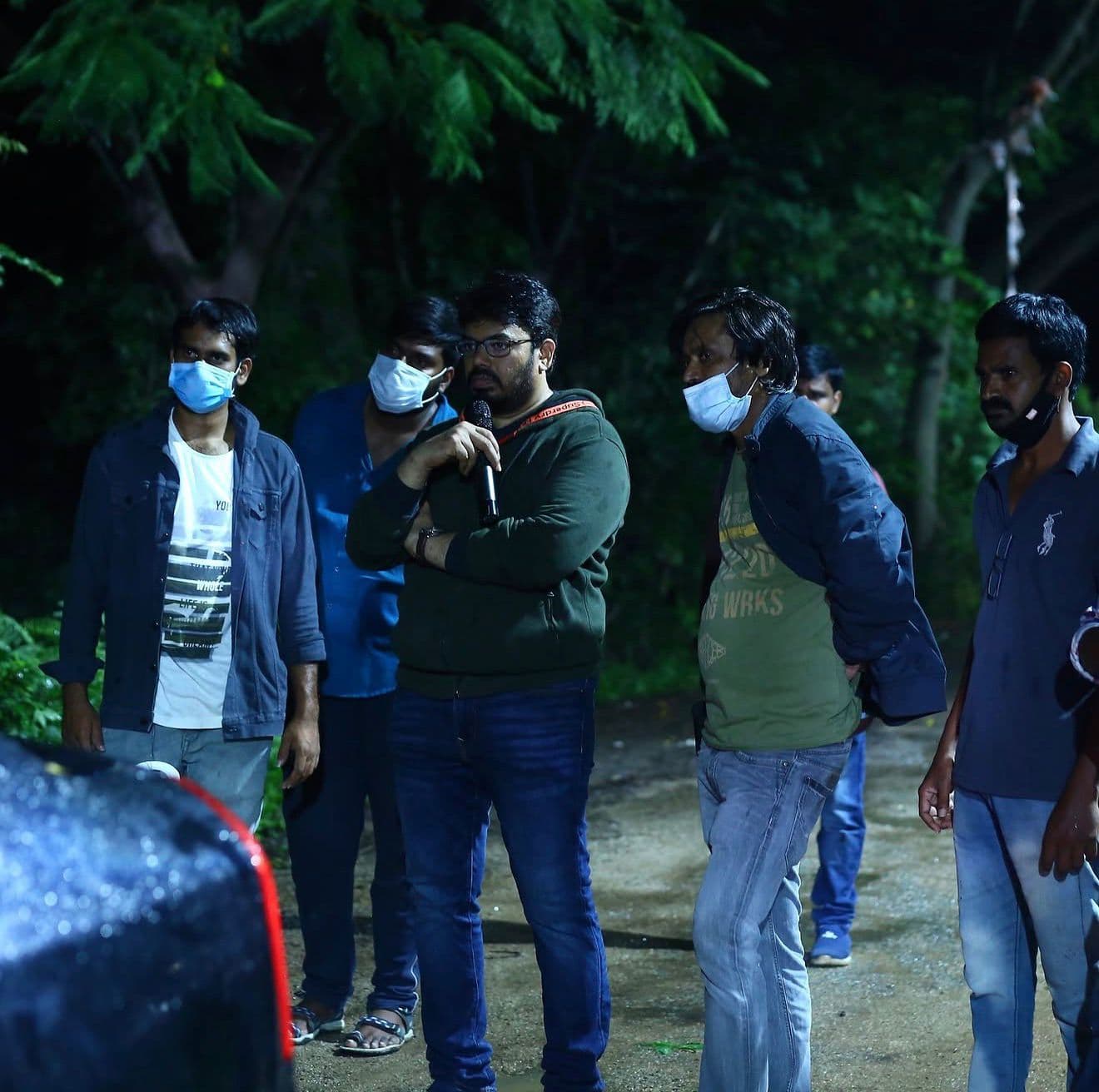 Crew standing together at night in a wooded area, one speaking into a microphone