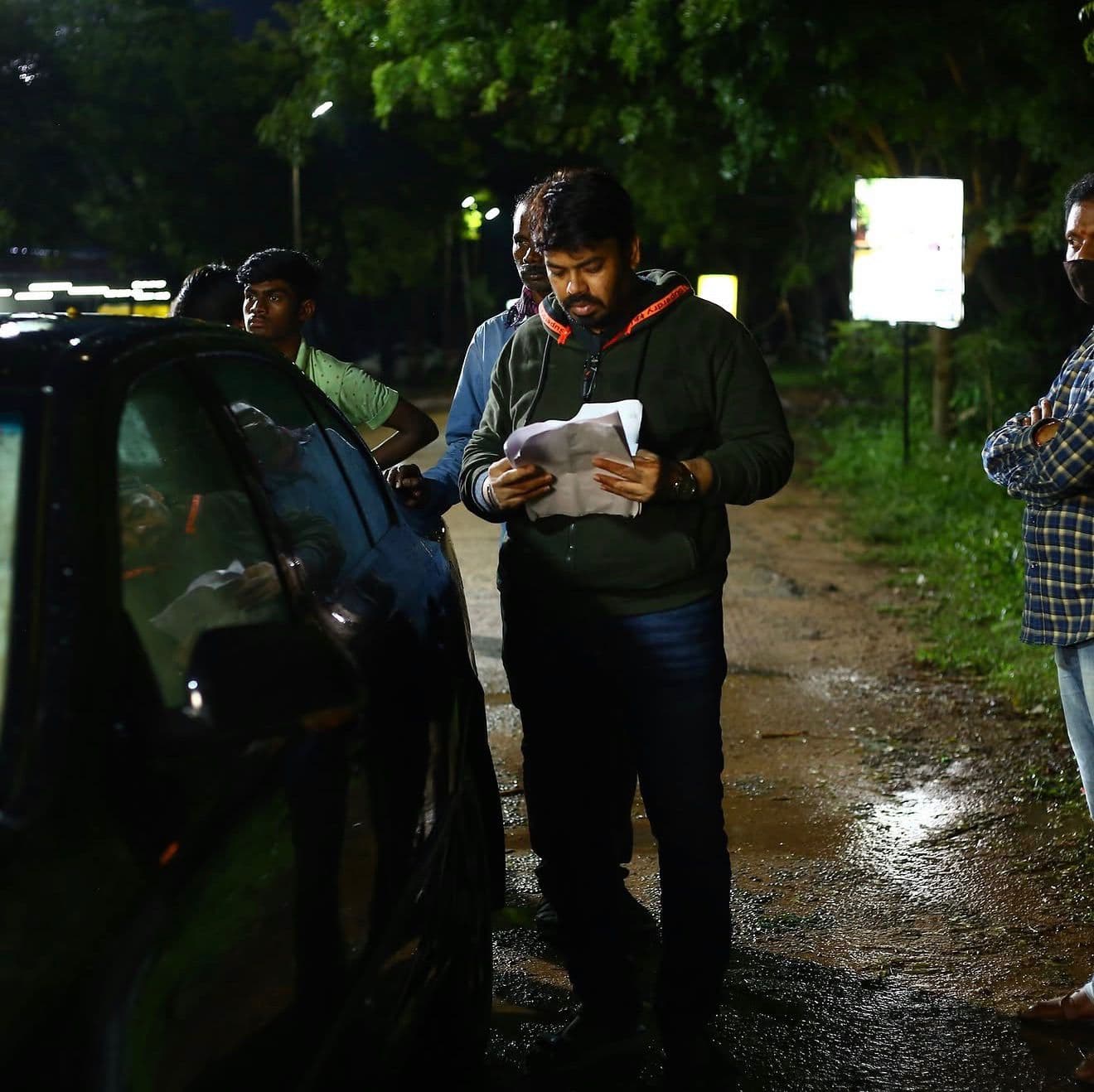 A crew member reviewing script pages beside a parked car at night, others looking on
