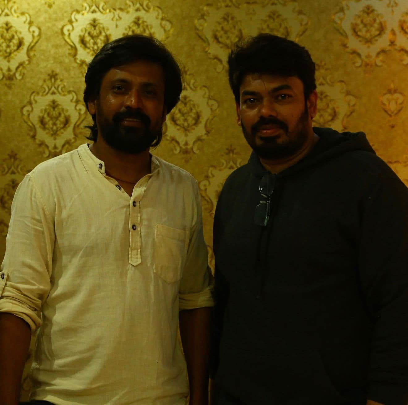 Two men posing together indoors against a patterned gold wallpaper