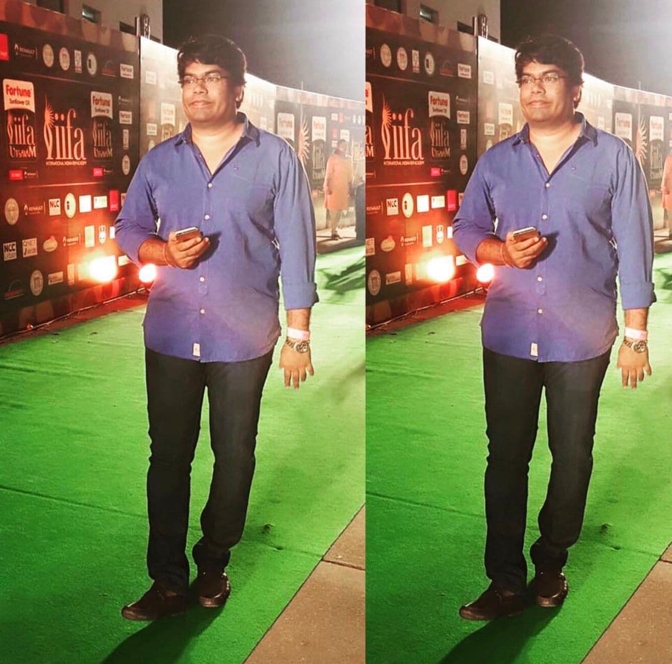 A man posing on a green event walkway in front of an IIFA step-and-repeat backdrop, holding a phone