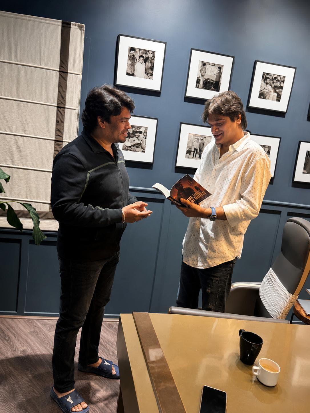 Two men in an office lined with black-and-white framed photographs, one holding an open paperback book