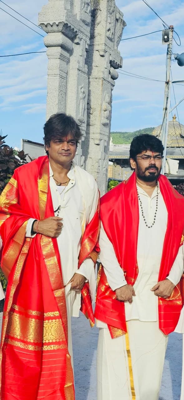 Two men wearing matching red and gold silk shawls over white traditional attire, standing beside a carved stone temple pillar