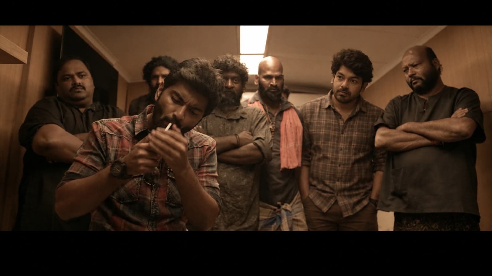 A cinematic film still of a man lighting a cigarette in a dimly lit hallway, surrounded by six other men standing with arms crossed