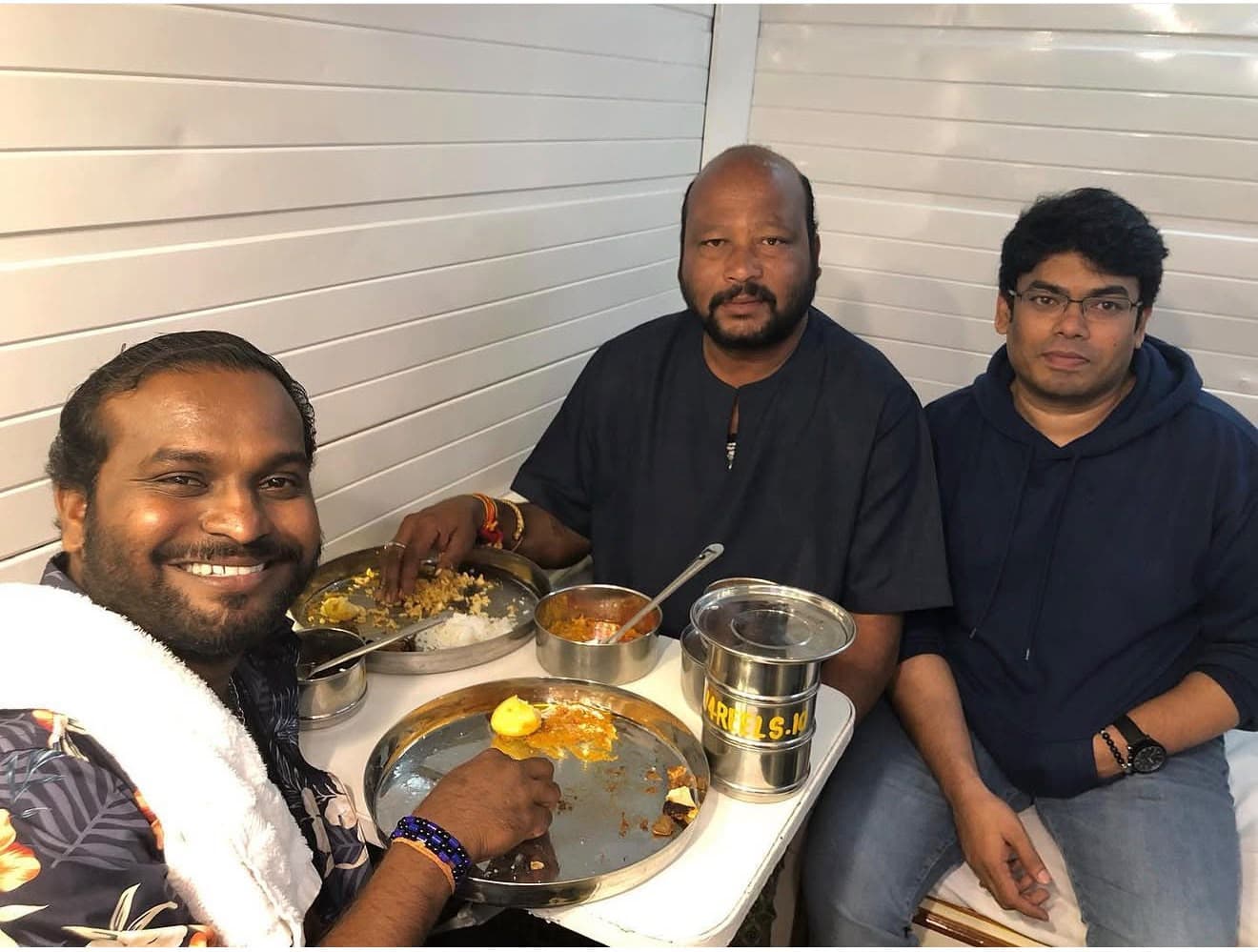 Three men sharing a meal on set, seated with steel plates and tiffin boxes of food