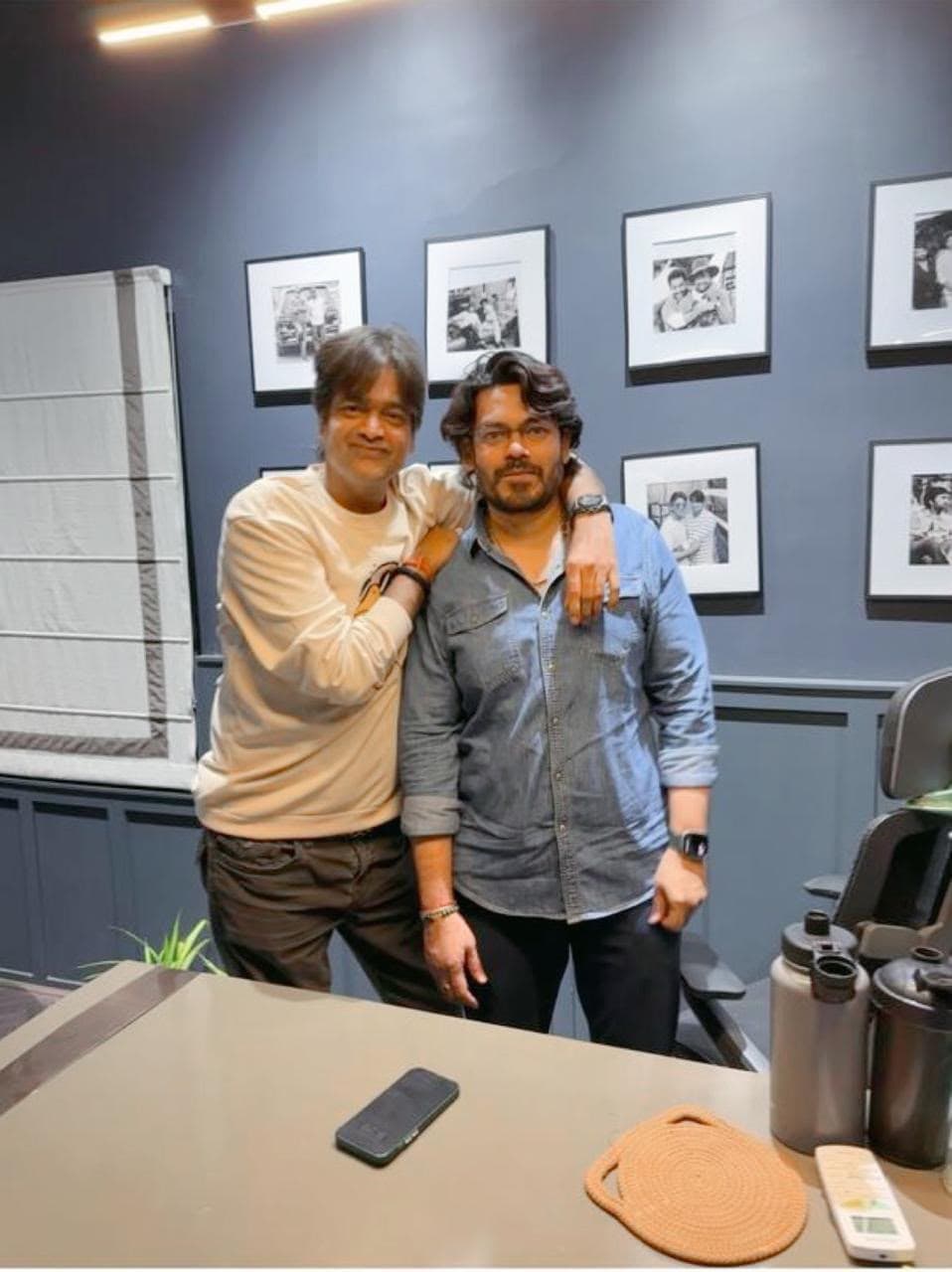 Two men posing arm in arm indoors, in front of a wall of framed black-and-white photographs