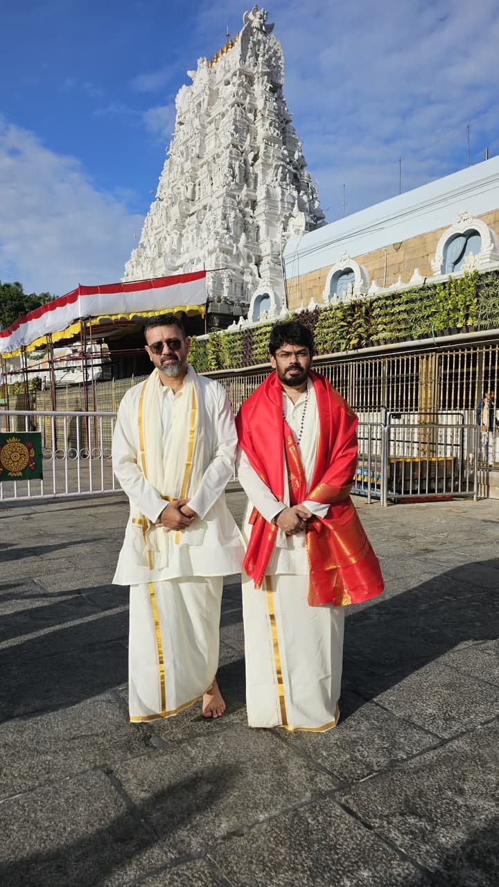 Two men in traditional white and gold-bordered dhotis, one wearing a red silk shawl, standing outside a South Indian temple with an ornately carved white tower
