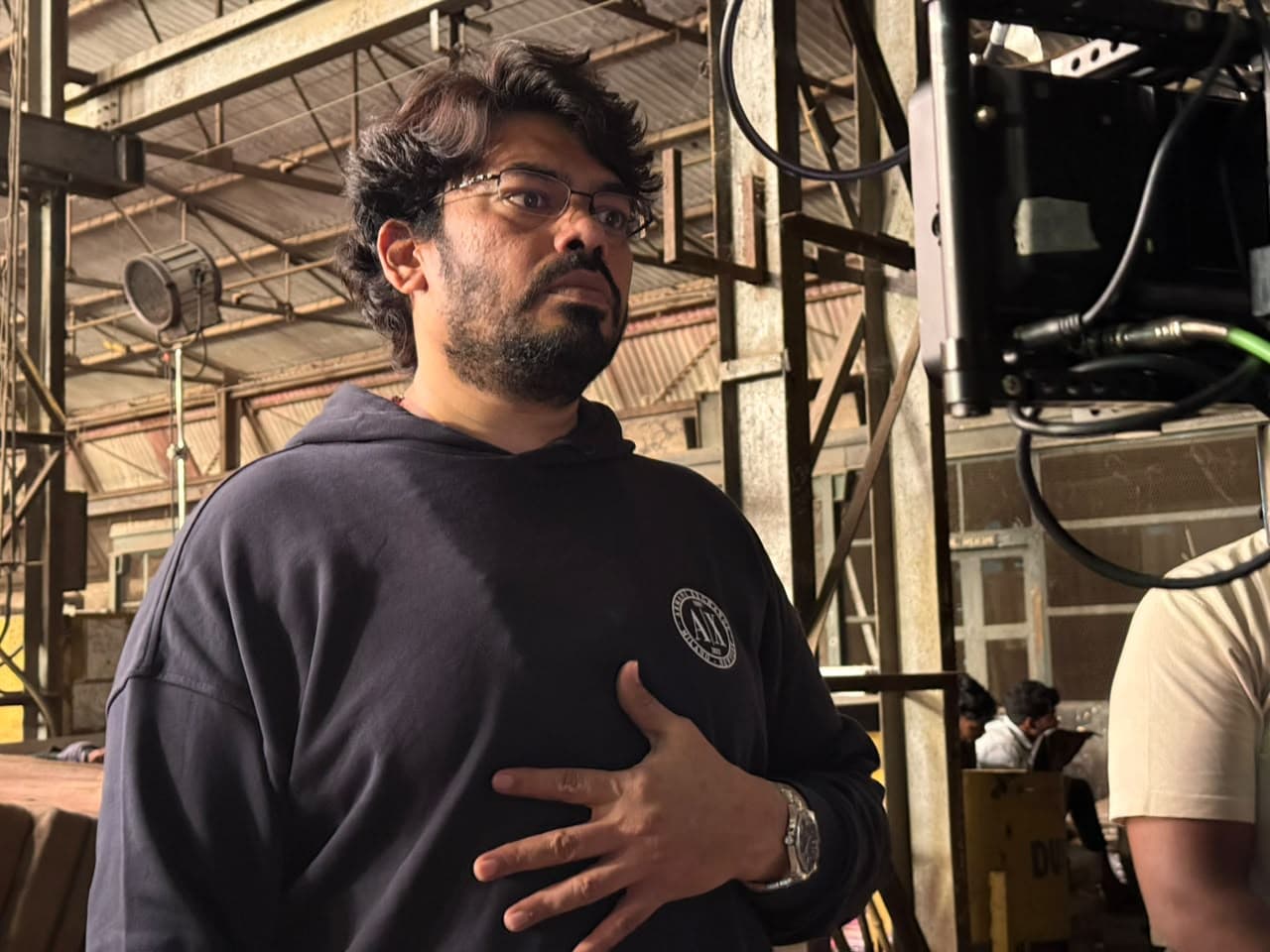 A man in a dark hoodie standing beside a camera monitor on a film set, looking off to the side