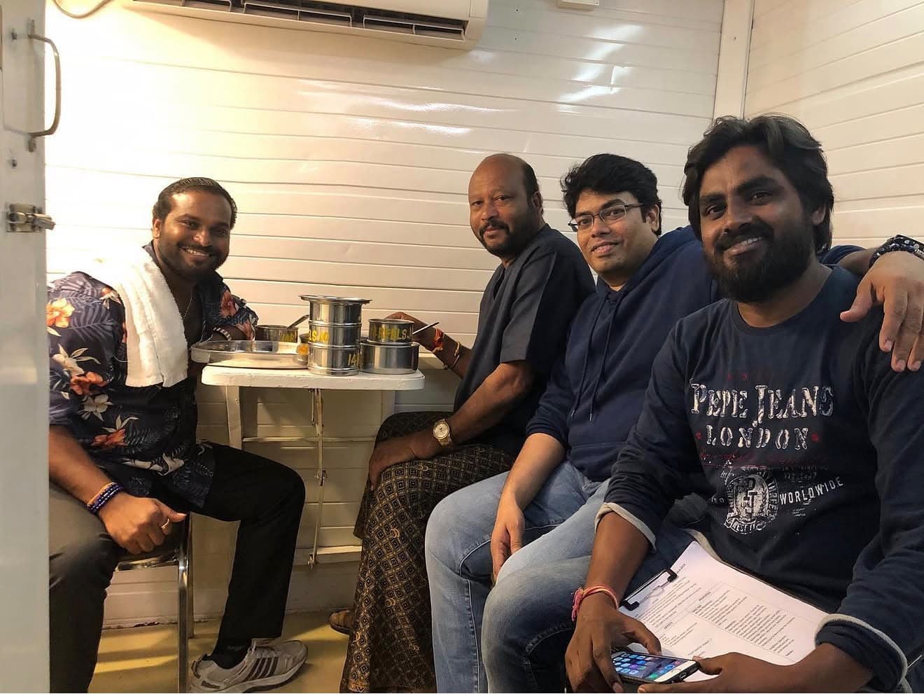 A group of four men smiling together on a break, seated beside a table with steel tiffin boxes