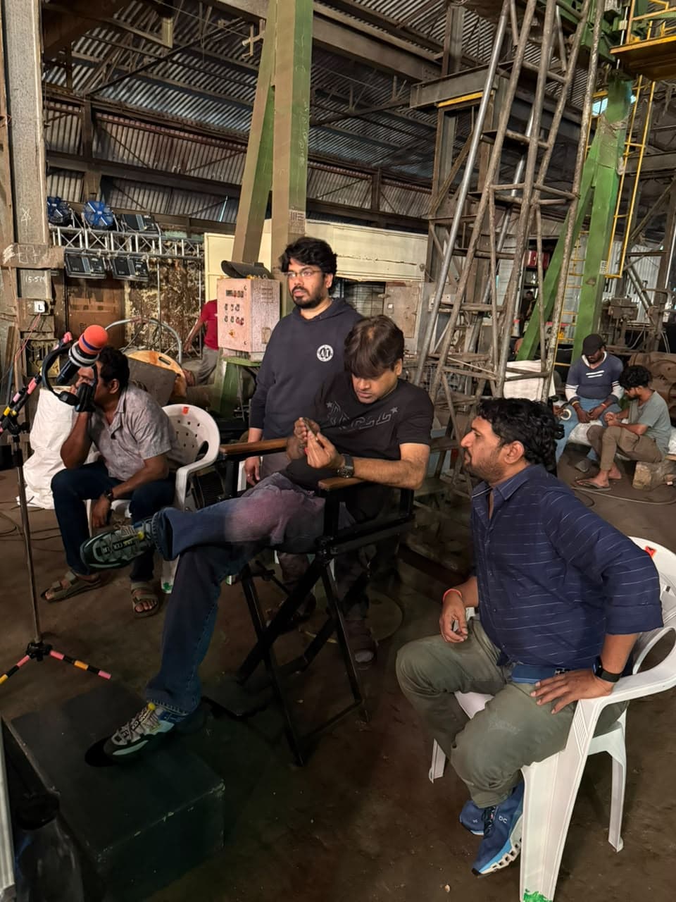 A group of film crew members on set, one seated in a director's chair, with a boom microphone operator nearby