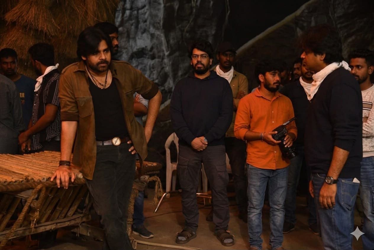 Behind-the-scenes photo of an actor in rugged costume on a rustic film set, with crew members standing nearby