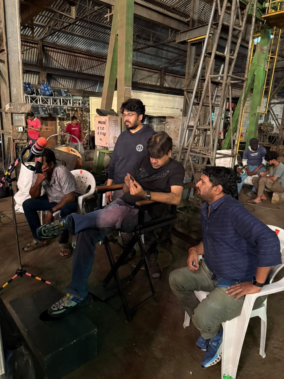 A group of film crew members gathered around a director's chair on a warehouse film set