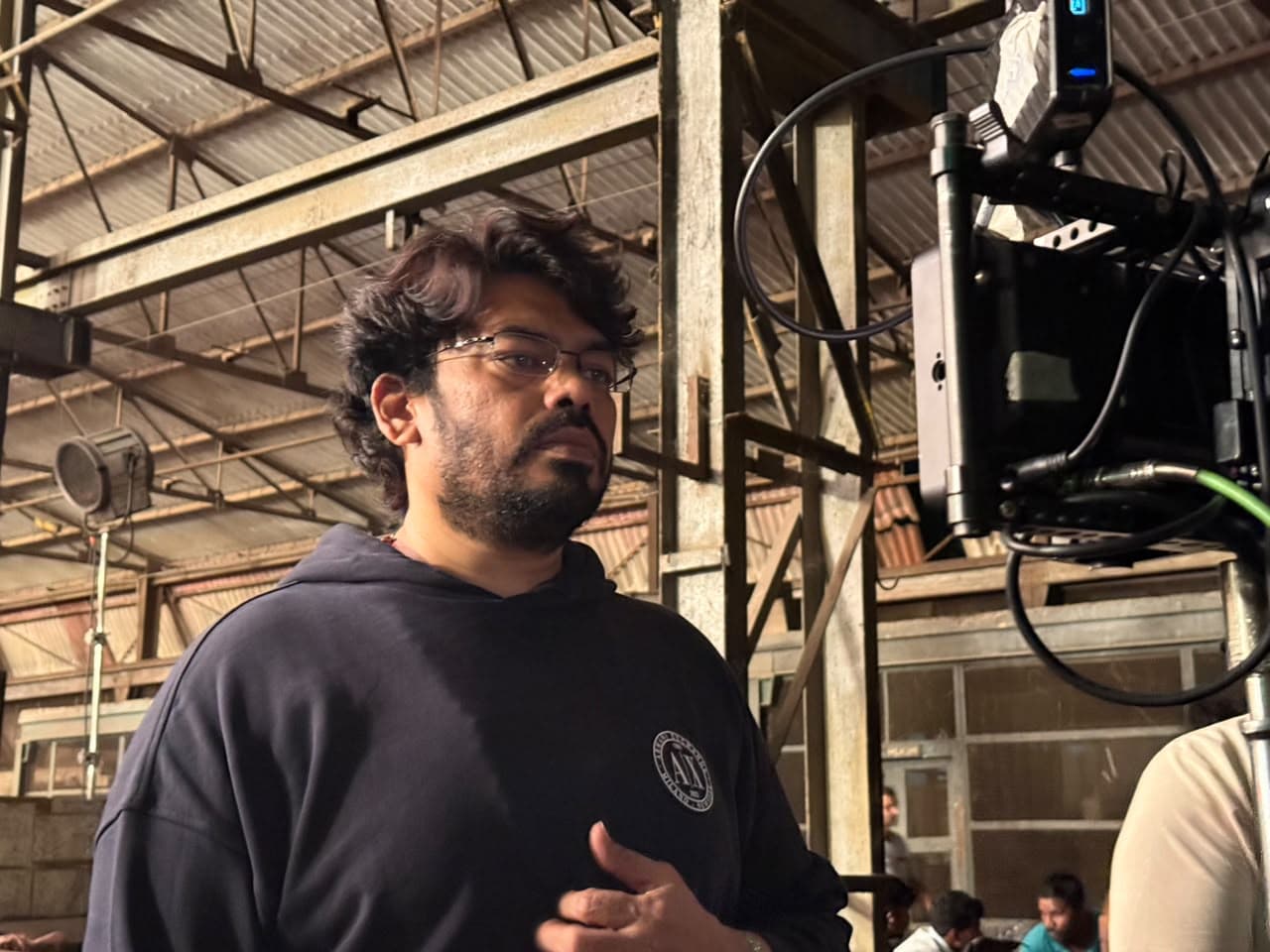 A man in a dark hoodie standing beside a camera monitor rig on a warehouse film set