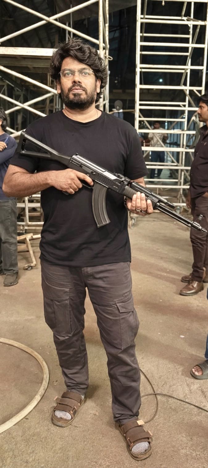 A man holding a prop rifle on a film set with metal scaffolding and crew members visible in the background