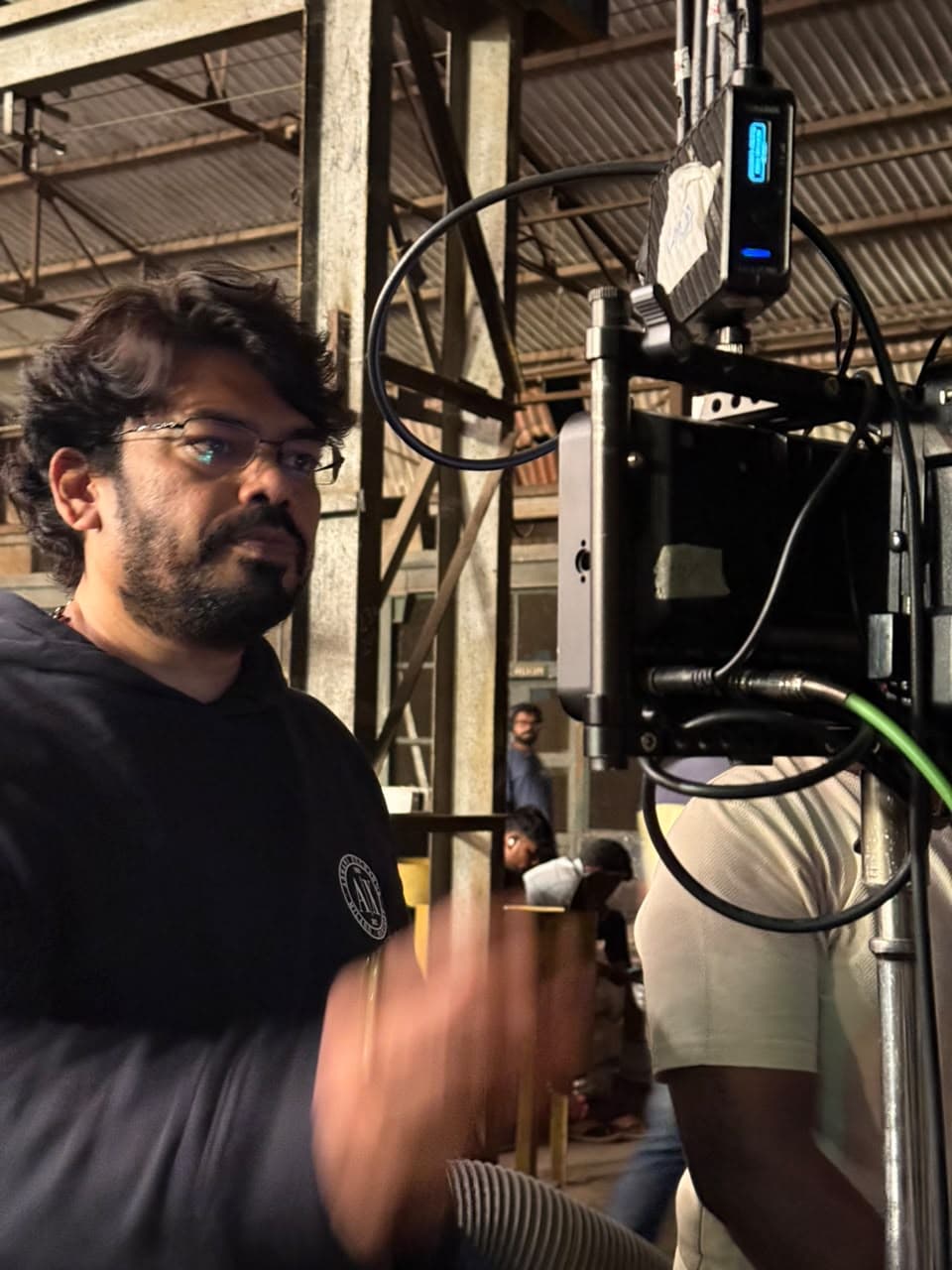 A man standing beside a camera monitor rig inside a warehouse-style film studio, gesturing while speaking